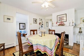 Une salle à manger avec une table recouverte d’une nappe à motifs, des chaises en bois, une cheminée blanche et des œuvres d’art encadrées accrochées aux murs.