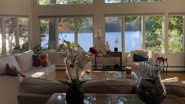A spacious living room with large windows overlooking a lake, furnished with a sofa, coffee table, and decorative items. - Waterfront, private dock, neighborhood beach (Saunderstown)