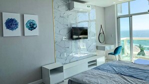Een moderne slaapkamer met een groot bed, een televisie die aan een marmeren muur is bevestigd, en uitzicht op de oceaan door grote ramen.