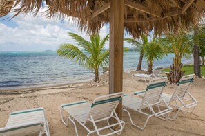 A beach with lounge chairs under a thatched roof, palm trees, and a view of the ocean.