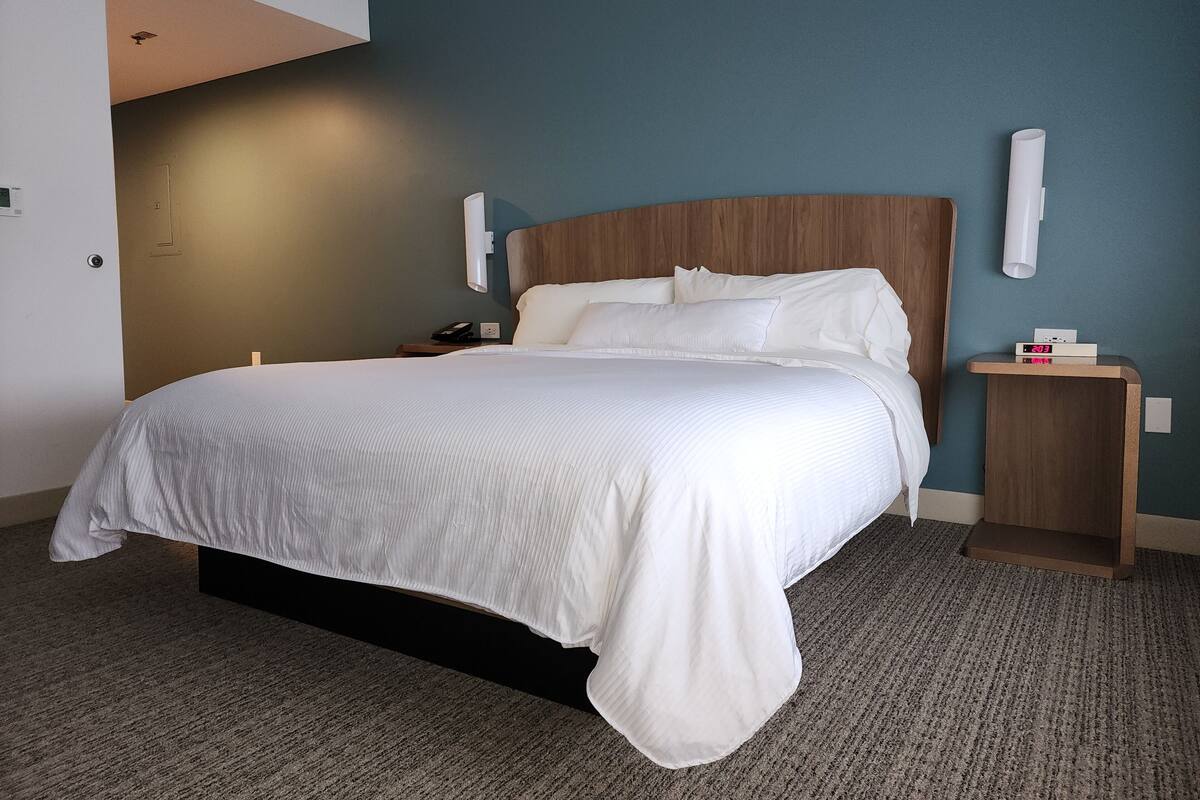 Ein Hotelzimmer mit einem großen Bett, einem Holzkopfstück, einem Nachttisch mit Telefon und einer an der Wand montierten Uhr.