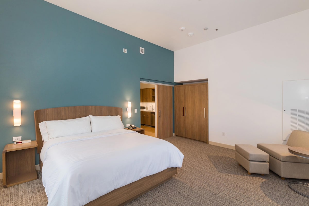 Ein modernes Hotelzimmer mit einem großen Bett, einem Nachttisch aus Holz, einer Sitzecke mit einem Sessel und einem Kleiderschrank.