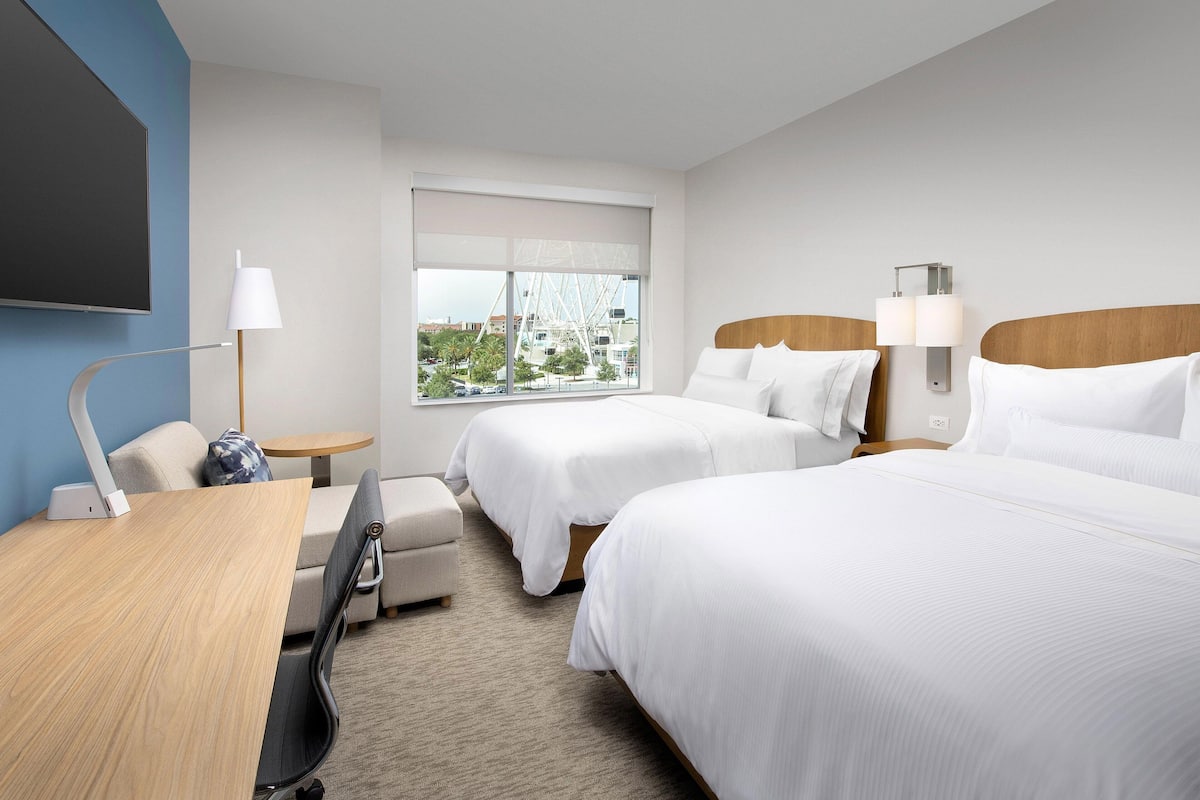 A hotel room with two beds, a desk, a chair, a lamp, and a window with a view of a Ferris wheel.