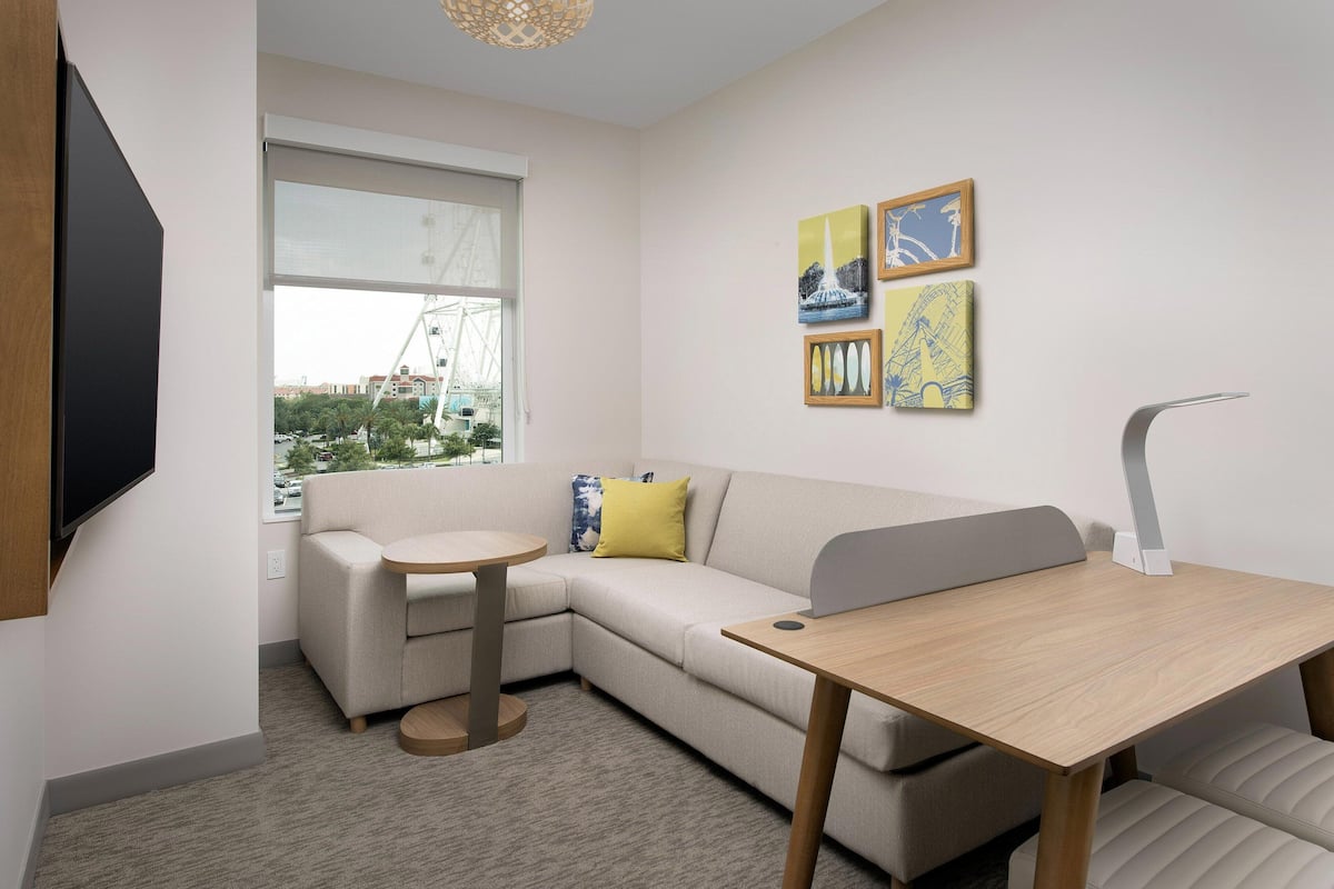 A modern room with a sofa, a small round table, and a desk. There is a window with a view of a building and a crane.