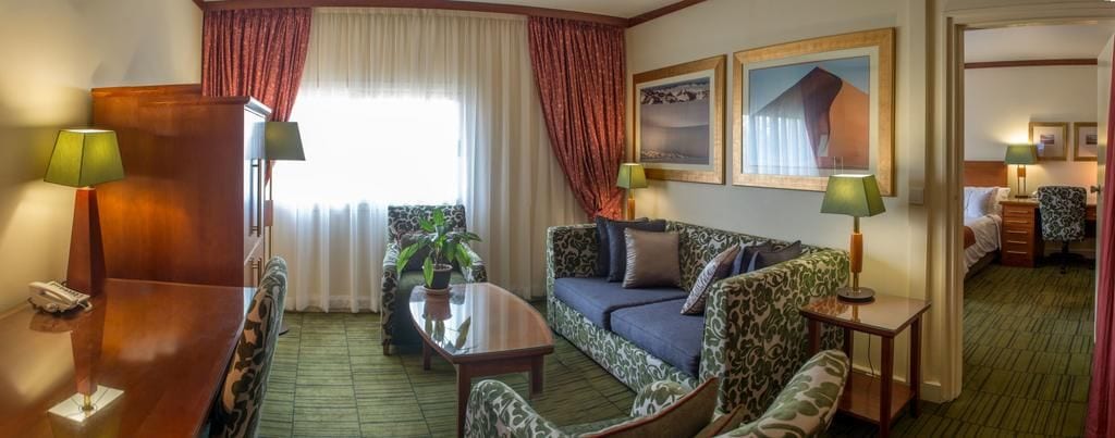 A hotel room with a sofa, a dining table, and a small coffee table. There are two lamps on the table and a plant on the sofa. The room has a window with curtains and a painting on the wall.