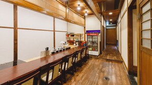 A traditional Japanese-style restaurant with a long wooden table and chairs, a kitchen area with a refrigerator, and a corridor leading to another room. - Couch Potato Hostel (Matsumoto)