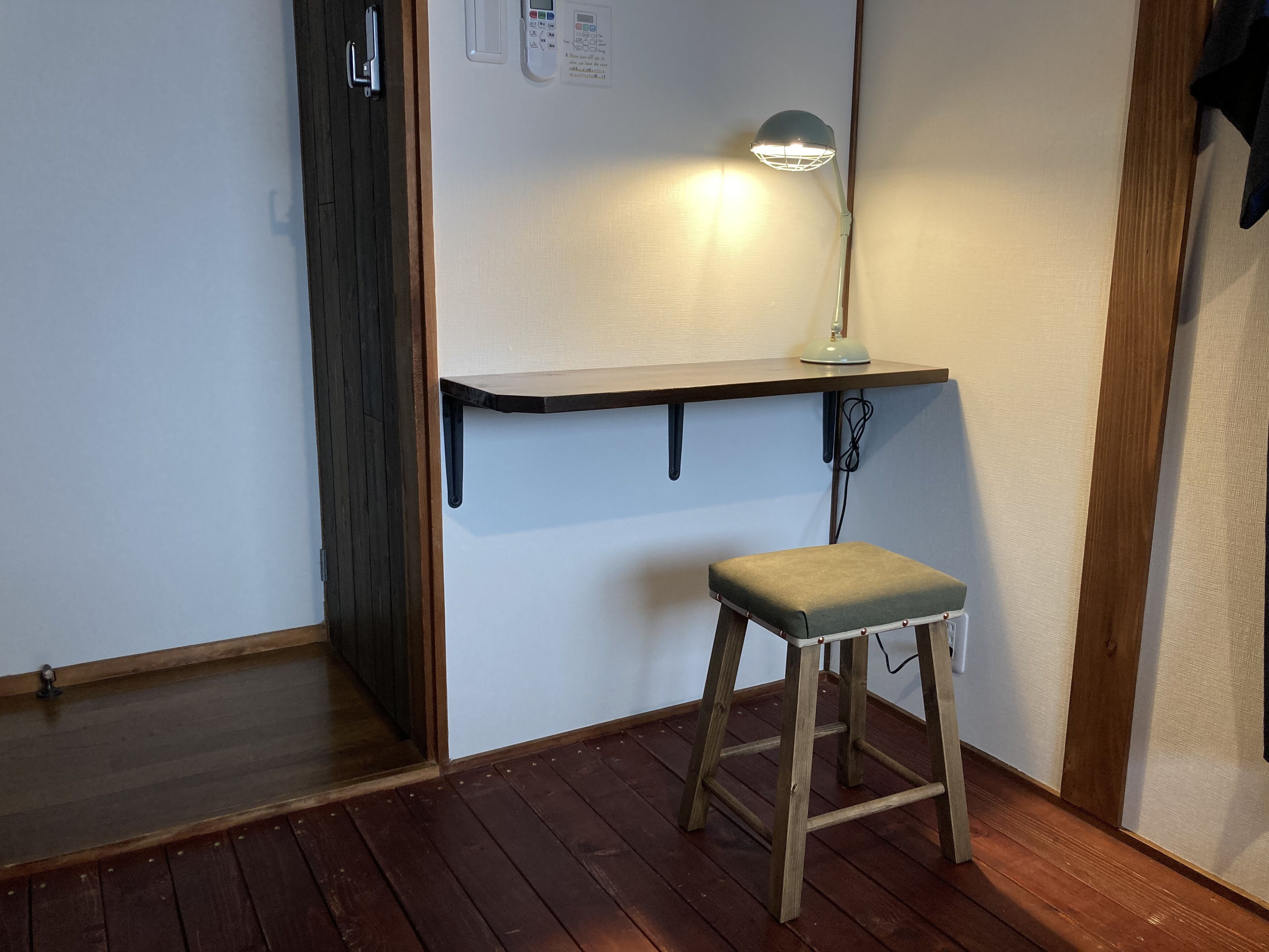 A small room with a wooden floor, a white wall, a wooden door, a small stool, and a desk with a lamp.