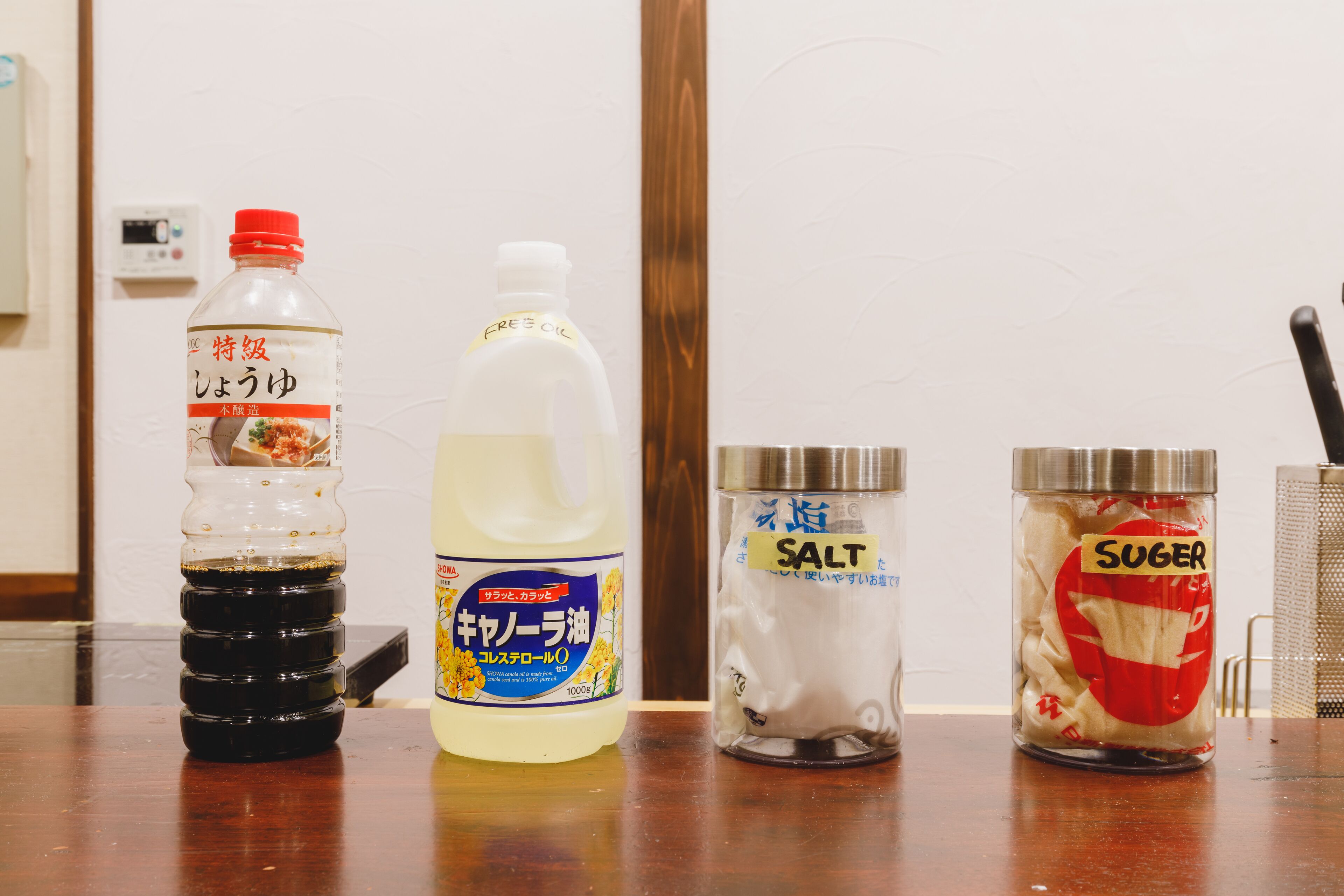 A table with three containers: a bottle of soy sauce, a bottle of cooking oil, and a jar of sugar.
