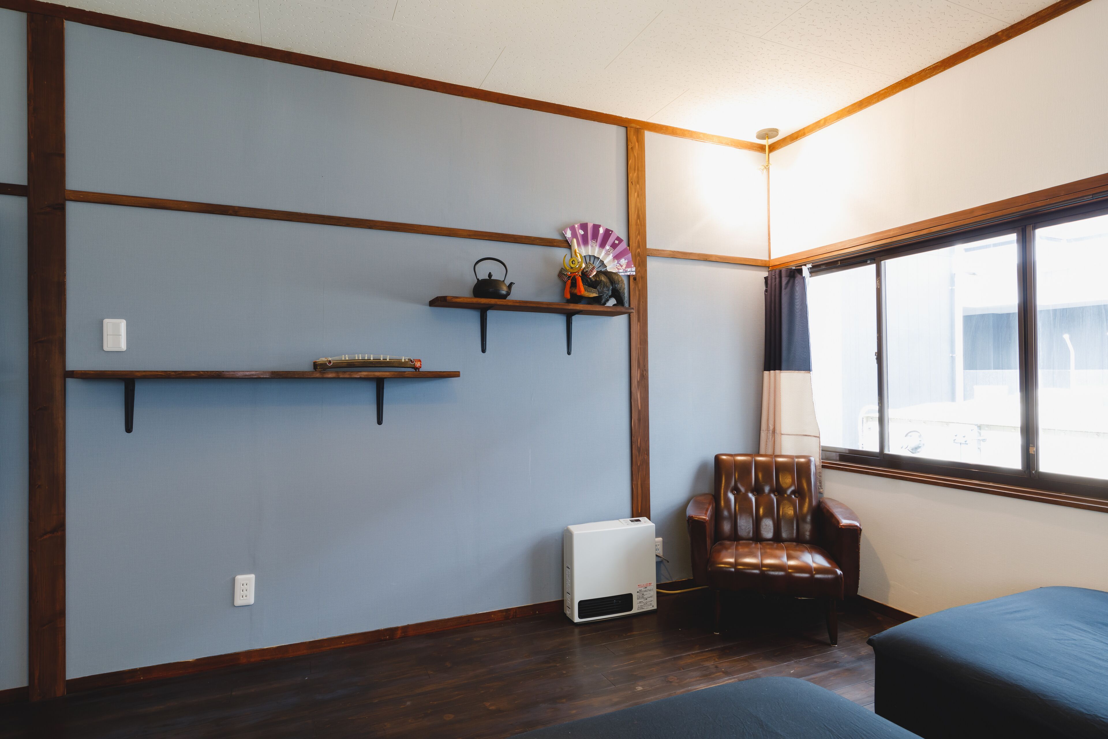 A room with a wooden floor, a brown leather chair, a bed, a window with a view, and a wall-mounted shelf with a kettle and a fan.