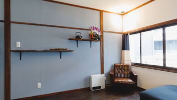 A room with a wooden floor, a brown leather chair, a bed, a window with a view, and a wall-mounted shelf with a kettle and a fan.