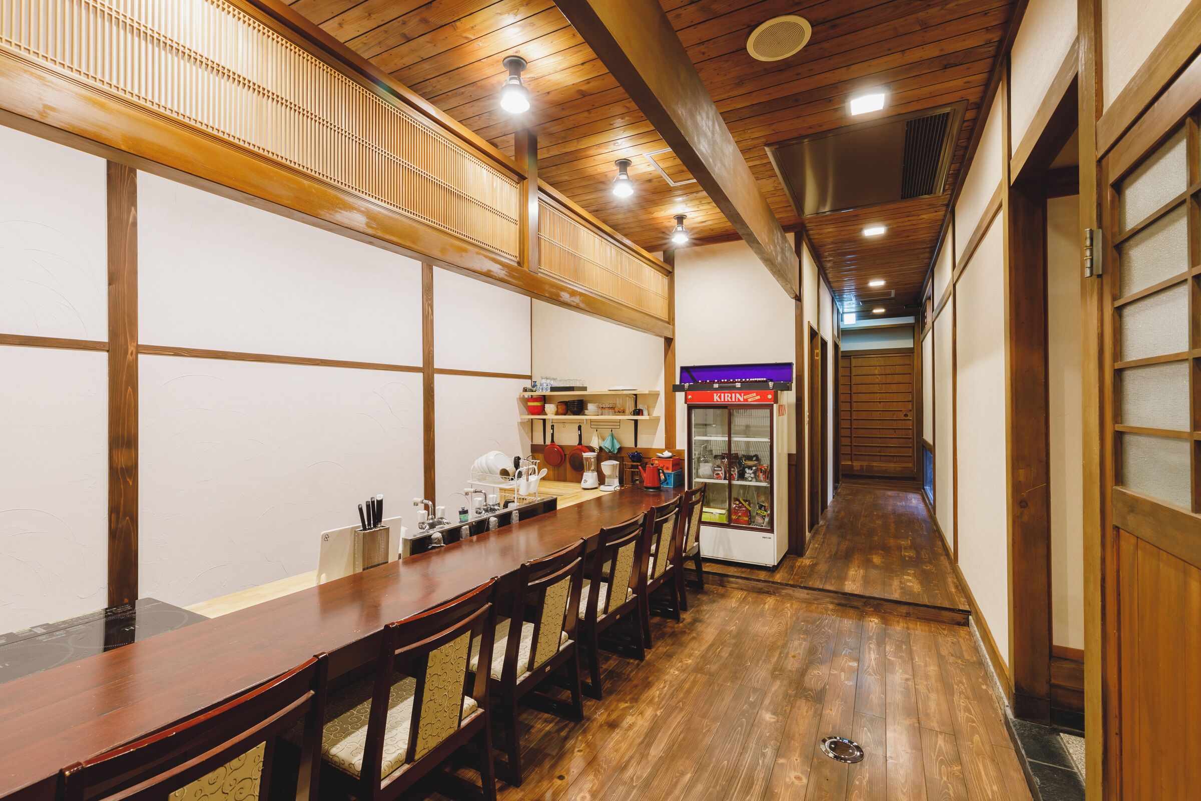 A traditional Japanese-style restaurant with a long wooden table and chairs, a kitchen area with a refrigerator, and a corridor leading to another room.