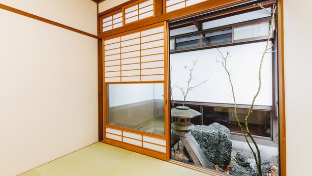 A traditional Japanese room with tatami flooring, shoji screens, and a view of a garden with a stone lantern and a rock. - Couch Potato Hostel (Matsumoto)