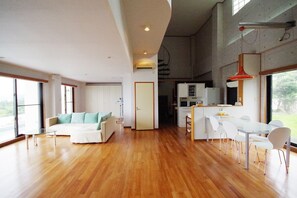 A spacious living area with a wooden floor, a white sofa with teal cushions, a dining table with chairs, and a kitchen area with white cabinets and a red pendant light. - John House (Ishigaki)