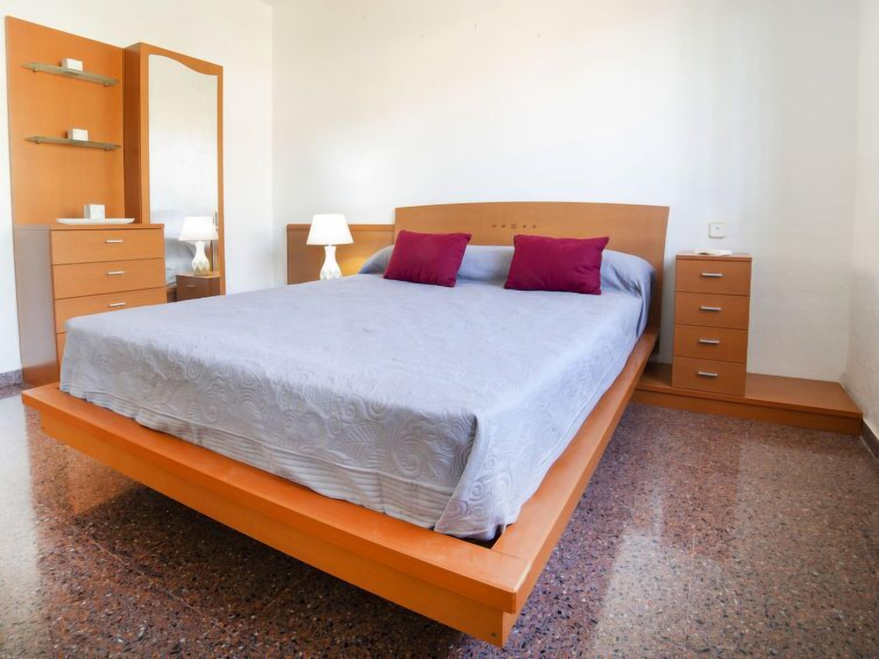 Un dormitori modern amb un llit gran, un panell de fusta al capçal, dues taules de nit, un prestatge i un mirall.