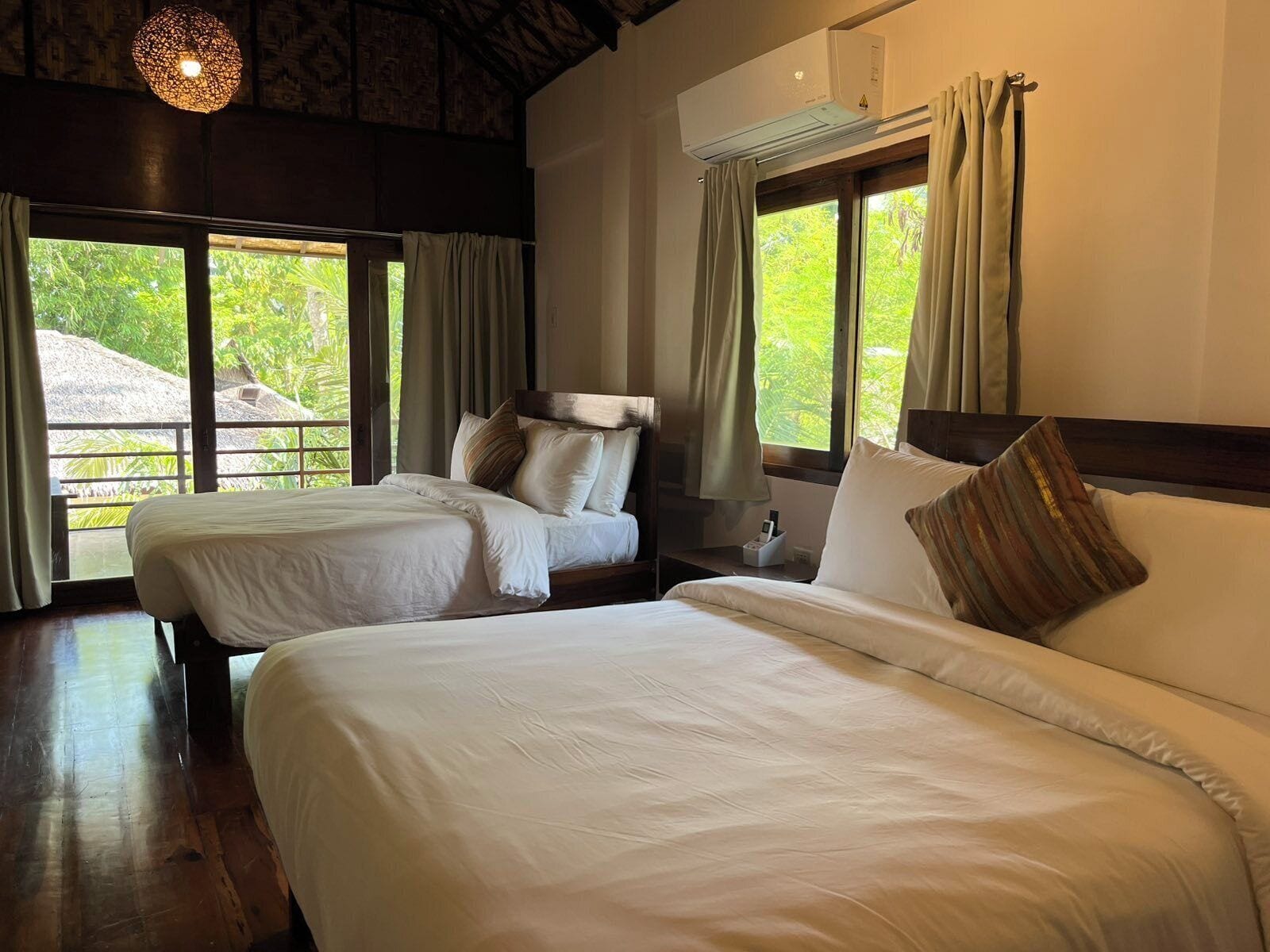 A bedroom with two beds, a ceiling fan, an air conditioning unit, and large windows with curtains.