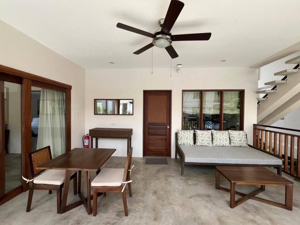 Una sala de estar con un ventilador de techo, un sofá con cojines estampados, una mesa de comedor de madera con sillas y una escalera.