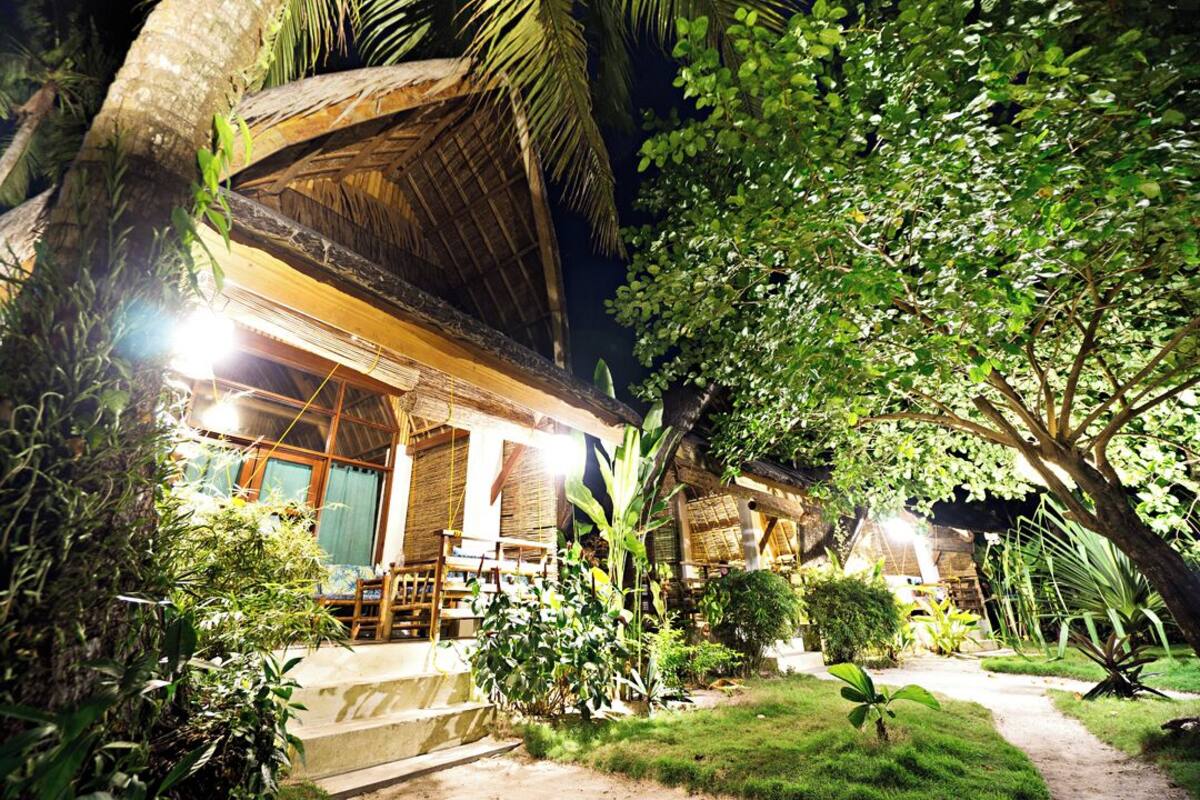 A rustic building with a thatched roof, surrounded by lush greenery and a well-lit pathway leading to the entrance.