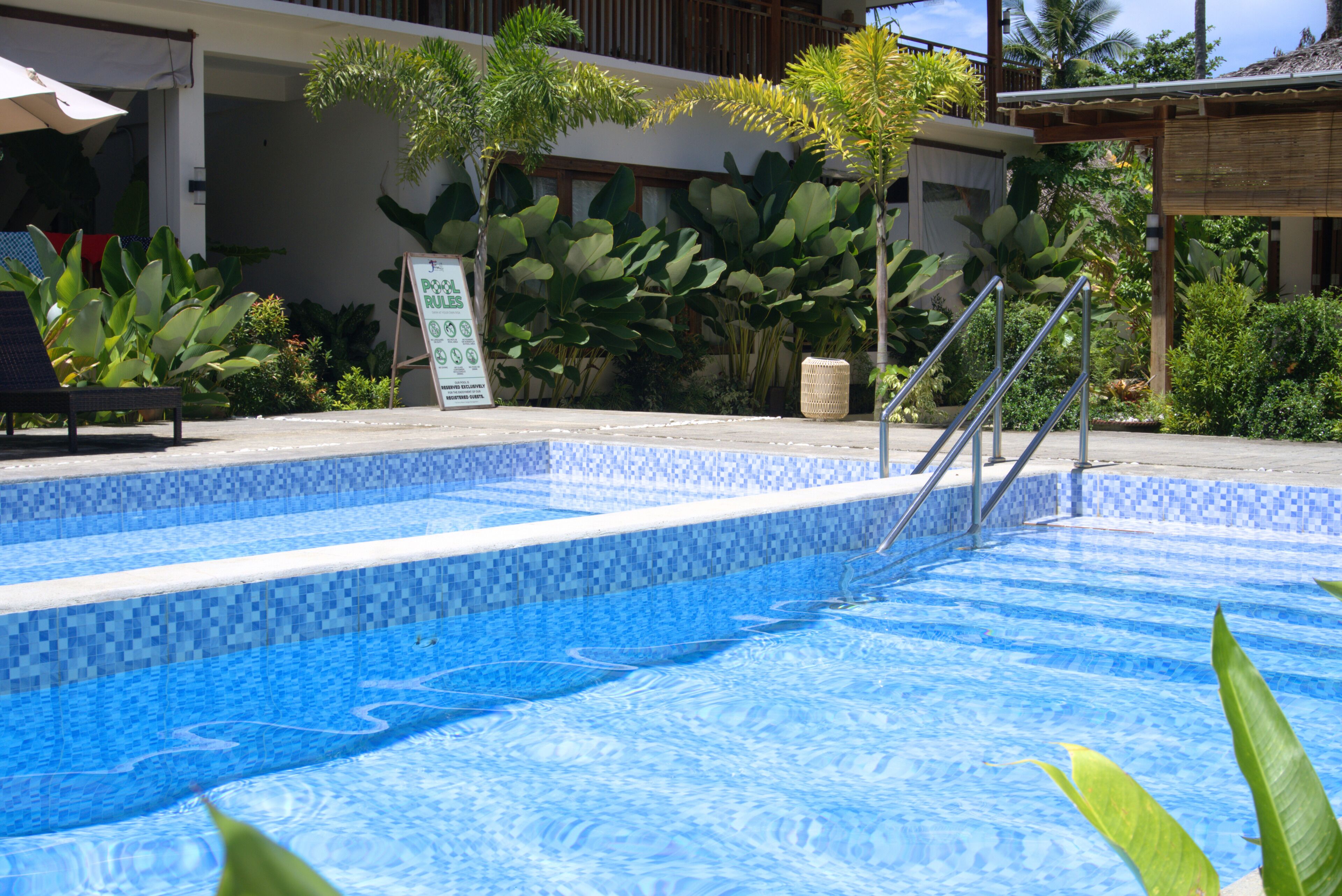 Una piscina amb vora de rajola, envoltada de plantes tropicals i un senyal que indica les normes de la piscina.