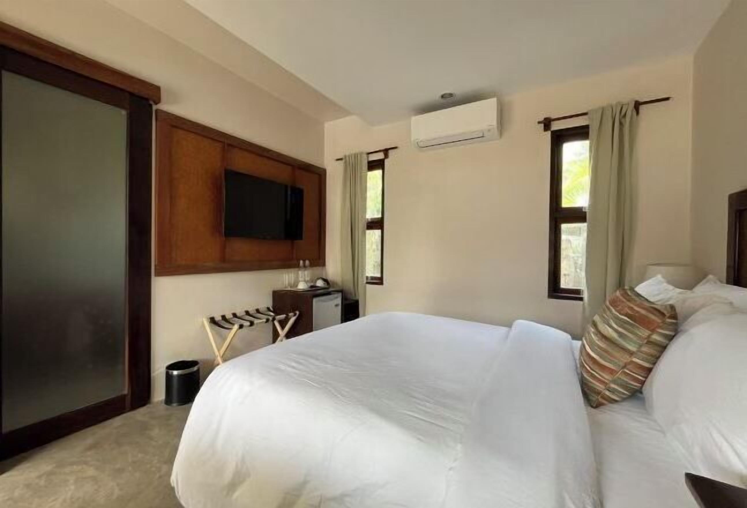 A hotel room with a large bed, a flat-screen TV mounted on the wall, a small desk with a lamp, and a window with curtains.