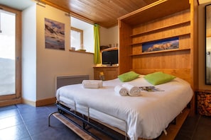 A hotel room with a bed, a television, wooden shelves, and a window with a view of mountains. - Apt Arsene1 Mont Blanc Views 3-min to Ski, Le Tour (Argentière), France (Le Tour (Argentière))