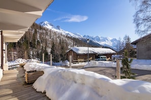 A wooden deck with snow accumulation, a wooden cabin with a snow-covered roof, and a mountain range in the background.