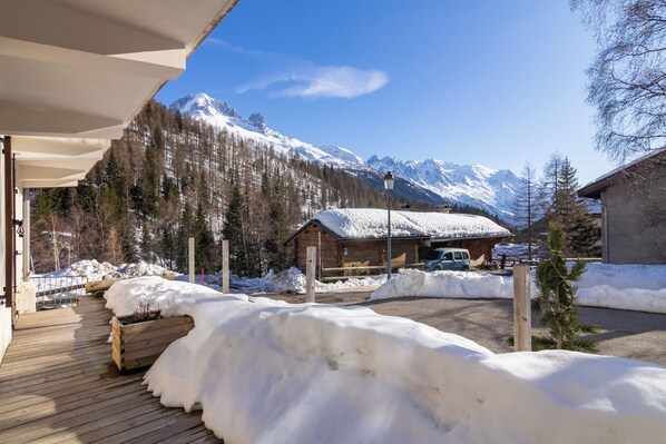 Eine Holzterrasse mit Schnee, eine Holzhütte mit verschneitem Dach und ein Gebirgszug im Hintergrund.