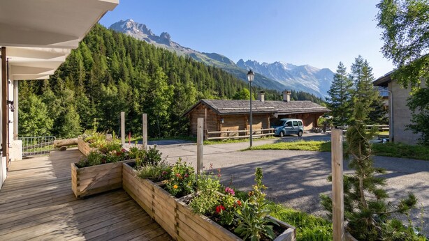 Un terrazza in legno con aiuole rialzate, vista sulle montagne, una piccola baita e un'auto parcheggiata.