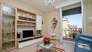 Un salon équipé d’un téléviseur, de quelques étagères ornées d’objets décoratifs, d’un vase avec des fleurs, d’un canapé et d’un balcon offrant une vue agréable.
