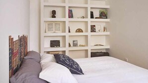 Une chambre à coucher comprenant un lit, une étagère blanche sur laquelle sont disposés divers objets, et un mur orné d’un panneau décoratif.