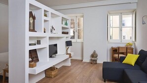 Un salon avec une bibliothèque blanche, un canapé bleu foncé agrémenté d’un coussin jaune, un plancher en bois et une fenêtre donnant sur des immeubles.