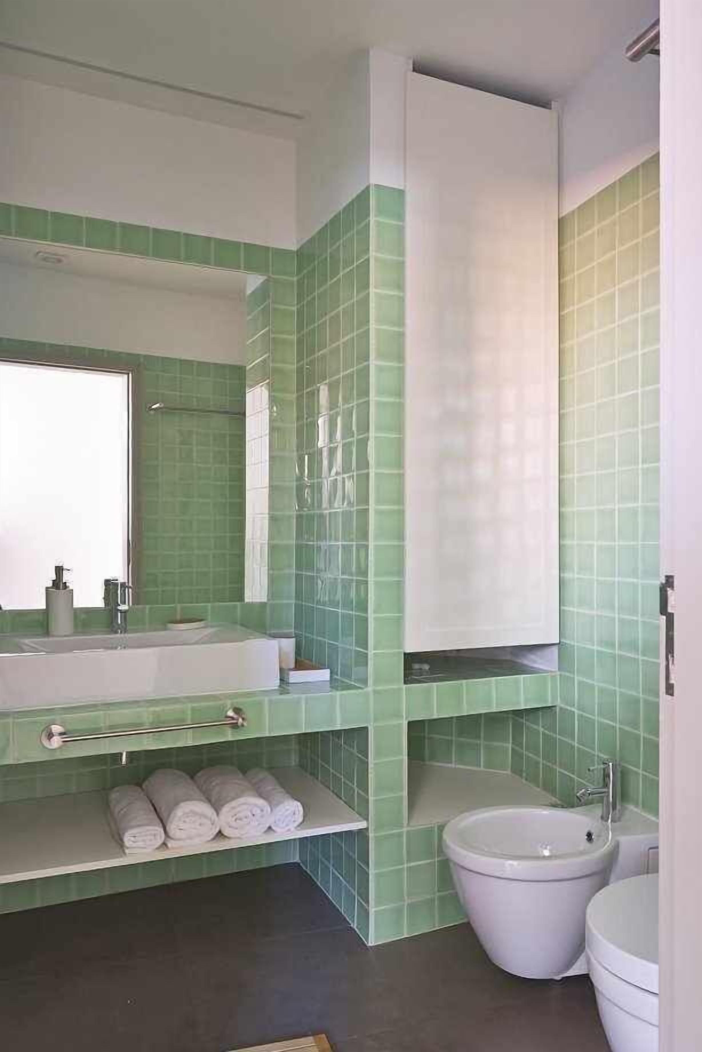 A bathroom with green tiled walls, a white sink, a toilet, and a large mirror.