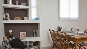 Ein Essbereich mit einem Holztisch, gedeckt für vier Personen, begleitet von zwei Holzstühlen. Eine weiße Schrankwand mit diversen Gegenständen steht links.
