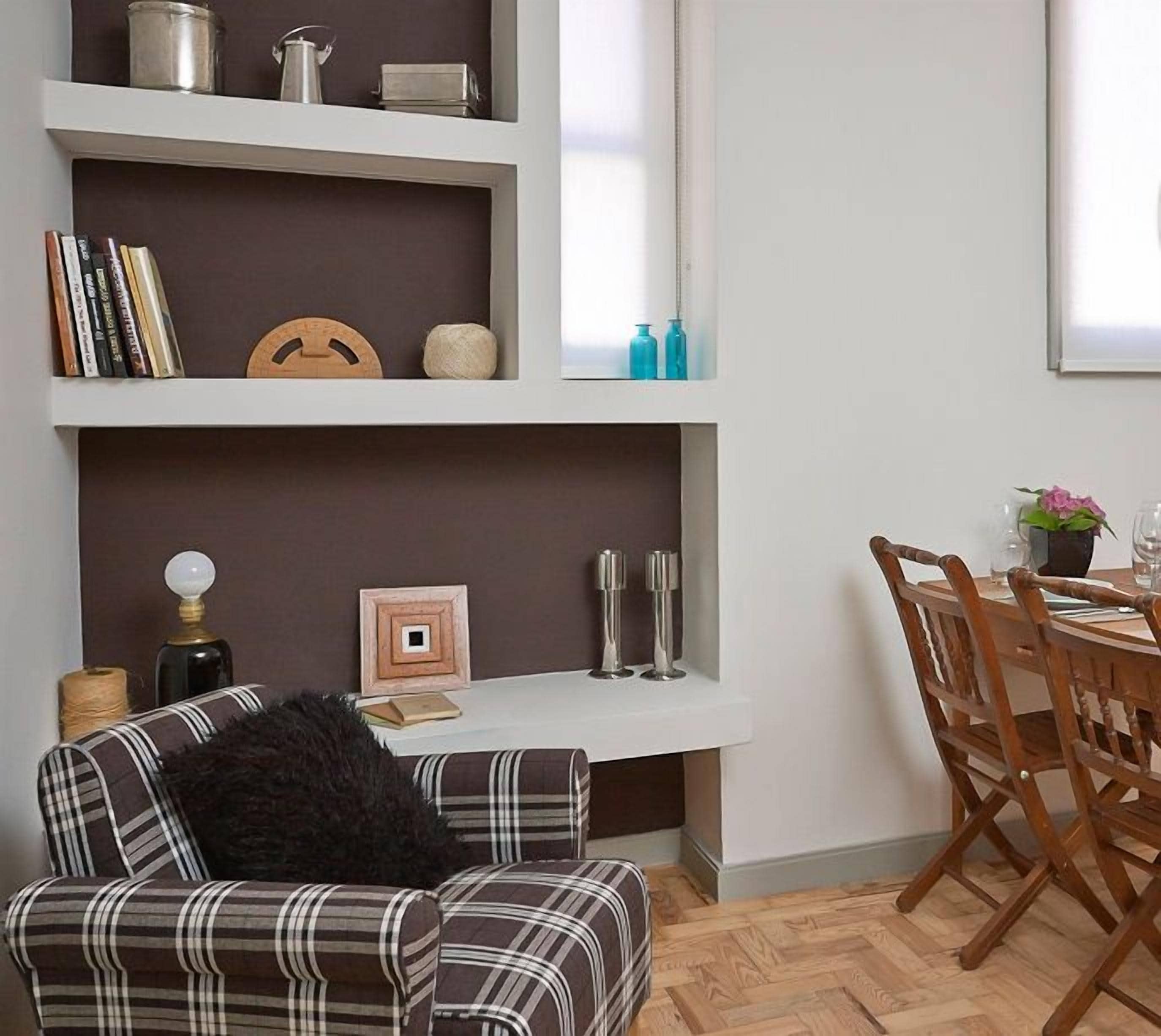 Een eetruimte met een houten tafel voor vier personen, omringd door houten stoelen. Links staat een boekenkast met diverse spullen erop, en erboven hangt een ingelijst schilderij.