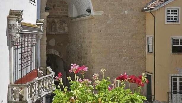 A stone building with a large arched entrance, a balcony with potted plants, and adjacent buildings with balconies. - Casas do Arco - Casa dos Mapas (Coimbra)