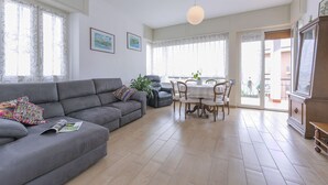 Un salón con un sofá gris, una mesa de comedor con sillas, una planta en maceta y vistas a través del ventanal.