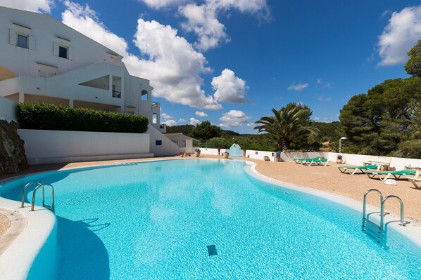 A swimming pool with a clear blue water, surrounded by a white building with balconies and a sandy beach area with lounge chairs. - Newly Renovated Apartment "Enzo" with Wi-Fi, Terrace & Pool (Son Parc)