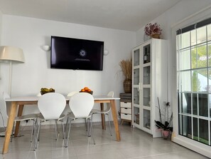 Uma sala de jantar moderna com televisão, cadeiras brancas, mesa de madeira e um armário com portas de vidro.
