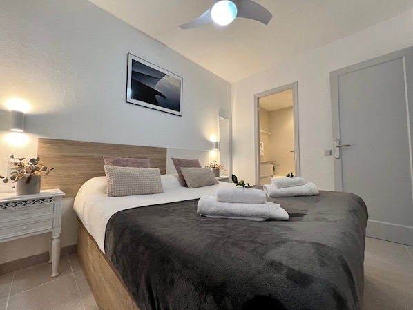 A modern hotel room with a large bed, bedside tables, a ceiling fan, a framed picture, and a bathroom visible through an open door.