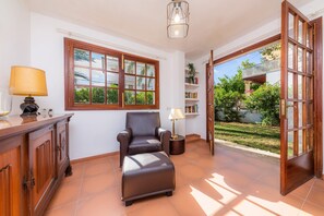 A room with a wooden cabinet, a leather chair, a small table with a lamp, a bookshelf, and a large window with a view of greenery. - Beautiful Villa "Beach Palmitos" with Wi-Fi, Balcony, Garden, Terraces & Pool (Can Picafort)