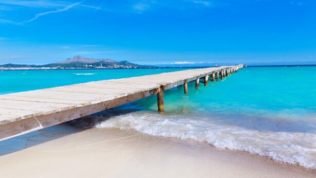 Une jetée en bois s’étendant dans une mer d’un bleu limpide, avec une plage de sable et une montagne en arrière-plan.