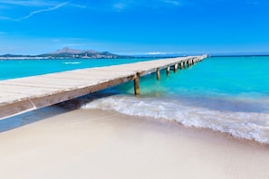 A wooden pier extending into a clear blue sea with a sandy beach and a mountain in the background.