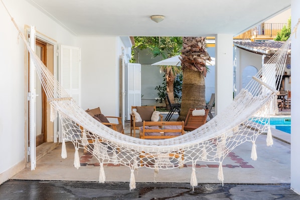 A hammock in a covered outdoor area with a pool and chairs.