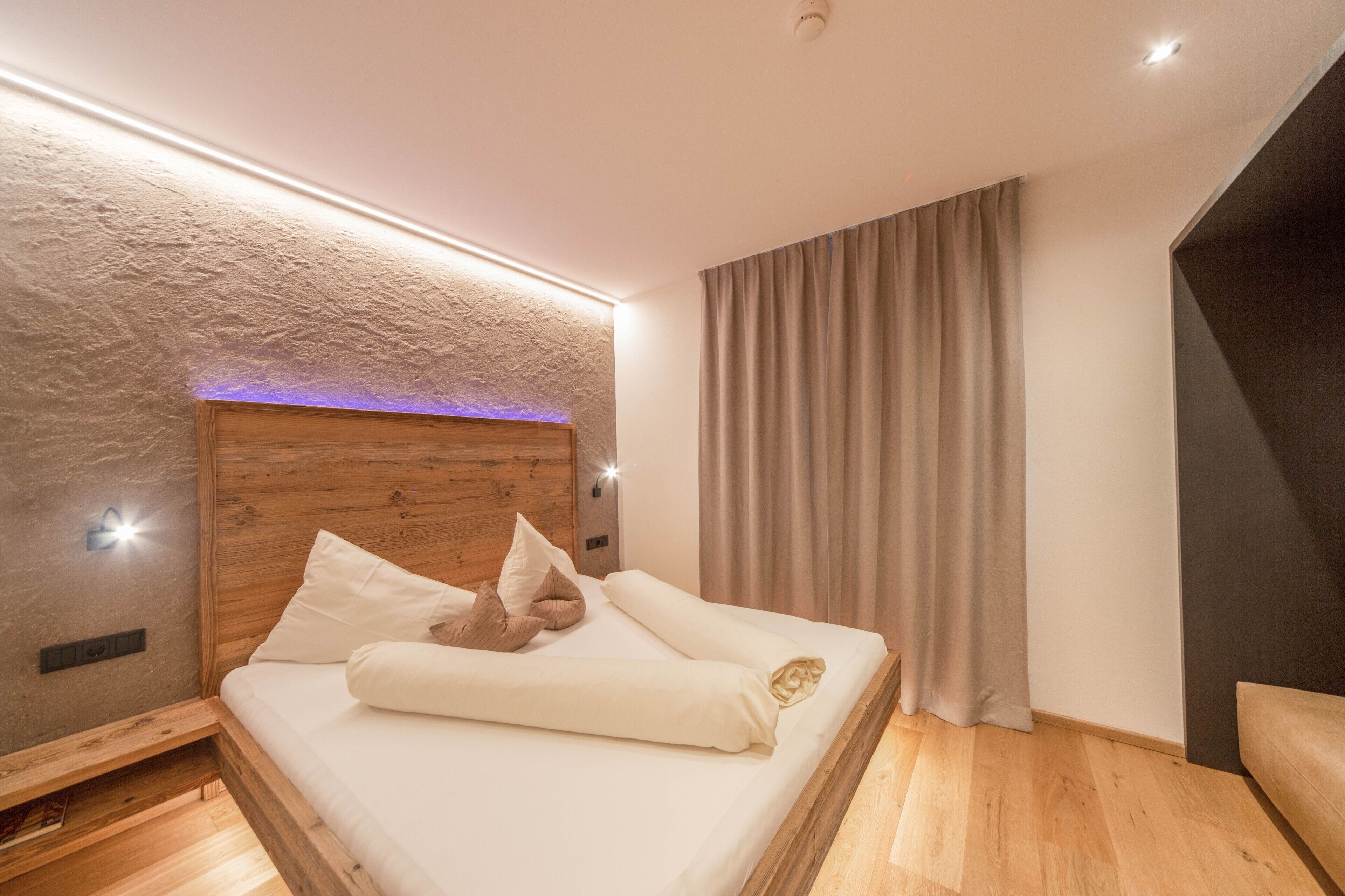 A modern bedroom with a wooden headboard, a bed with white linens, a built-in shelf, and a large window with curtains.