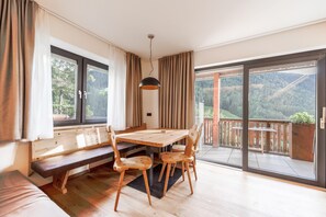 Ein Zimmer mit einem Holzesstisch, Stühlen, einer Bank und einer großen Schiebetür, die auf einen Balkon mit Blick auf eine bewaldete Hügellandschaft führt.