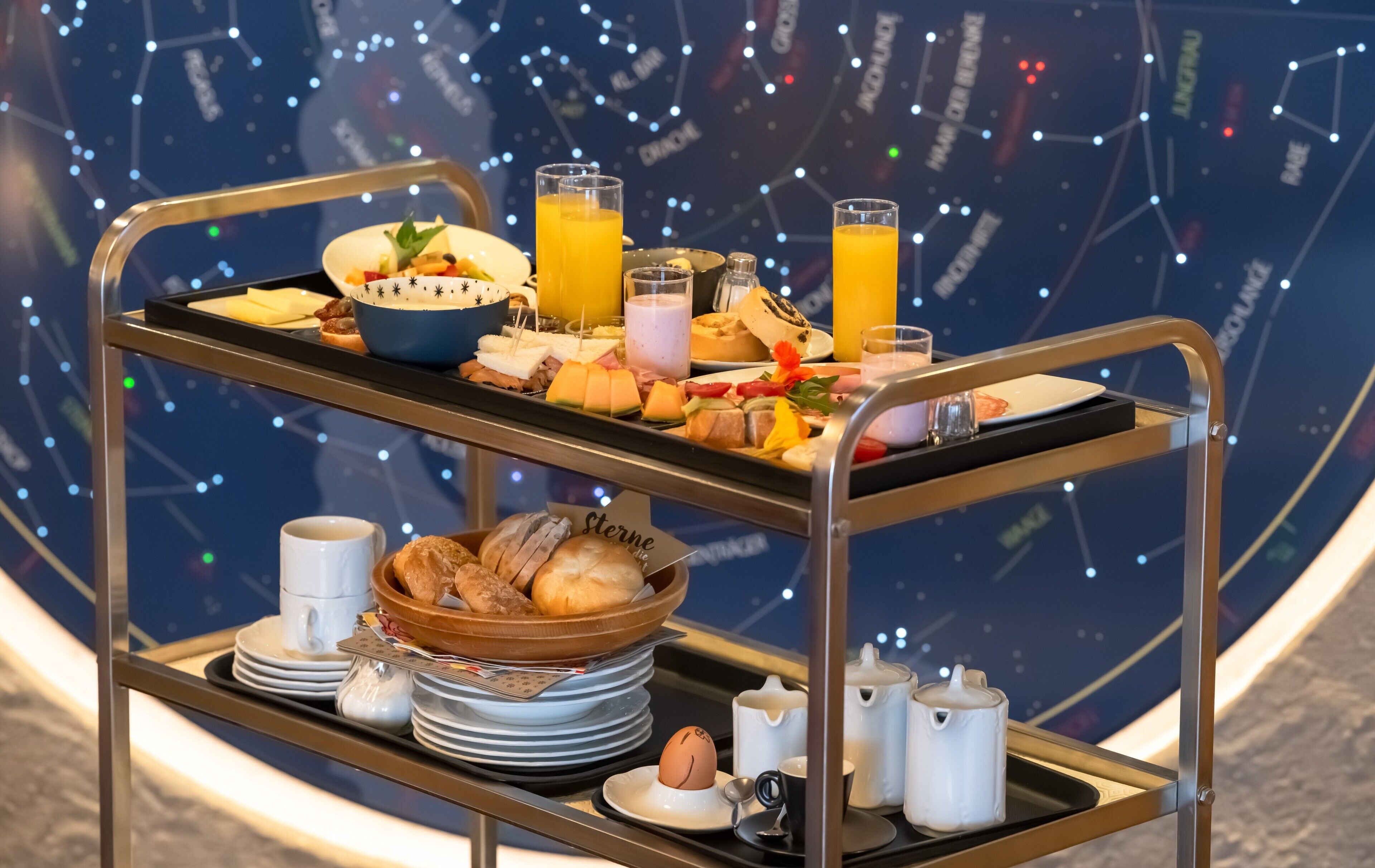 A breakfast trolley with various food items and drinks, a bowl of bread, plates, and cups, set against a starry background.