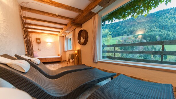 Une pièce avec un plafond en bois, un lit avec un oreiller, une chaise, une fenêtre donnant sur une forêt et une horloge murale.