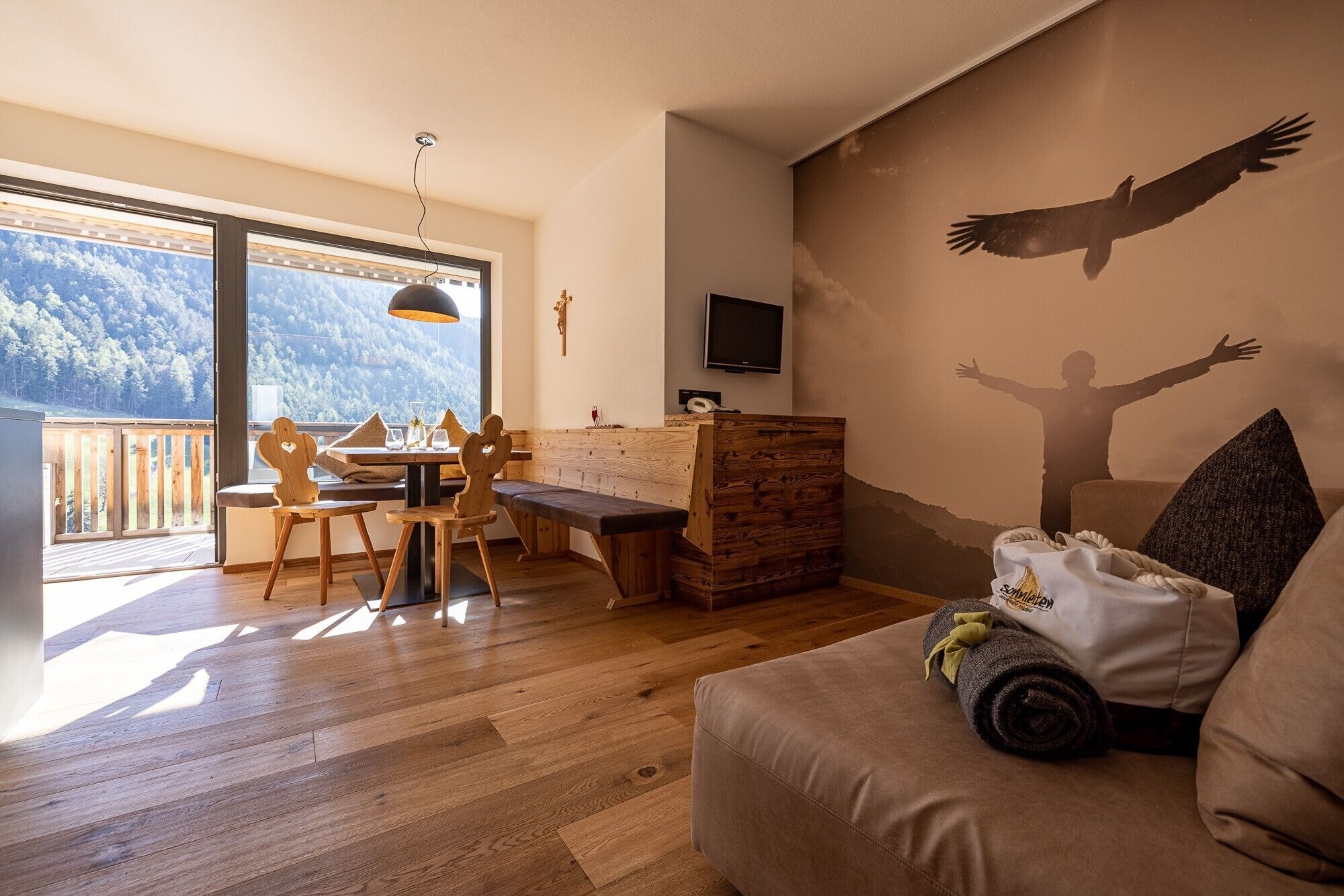 Ein Zimmer mit einem Holzes Esstisch, Stühlen, einer Couch, einem Fernseher und einem Wandgemälde einer Person mit ausgebreiteten Armen.