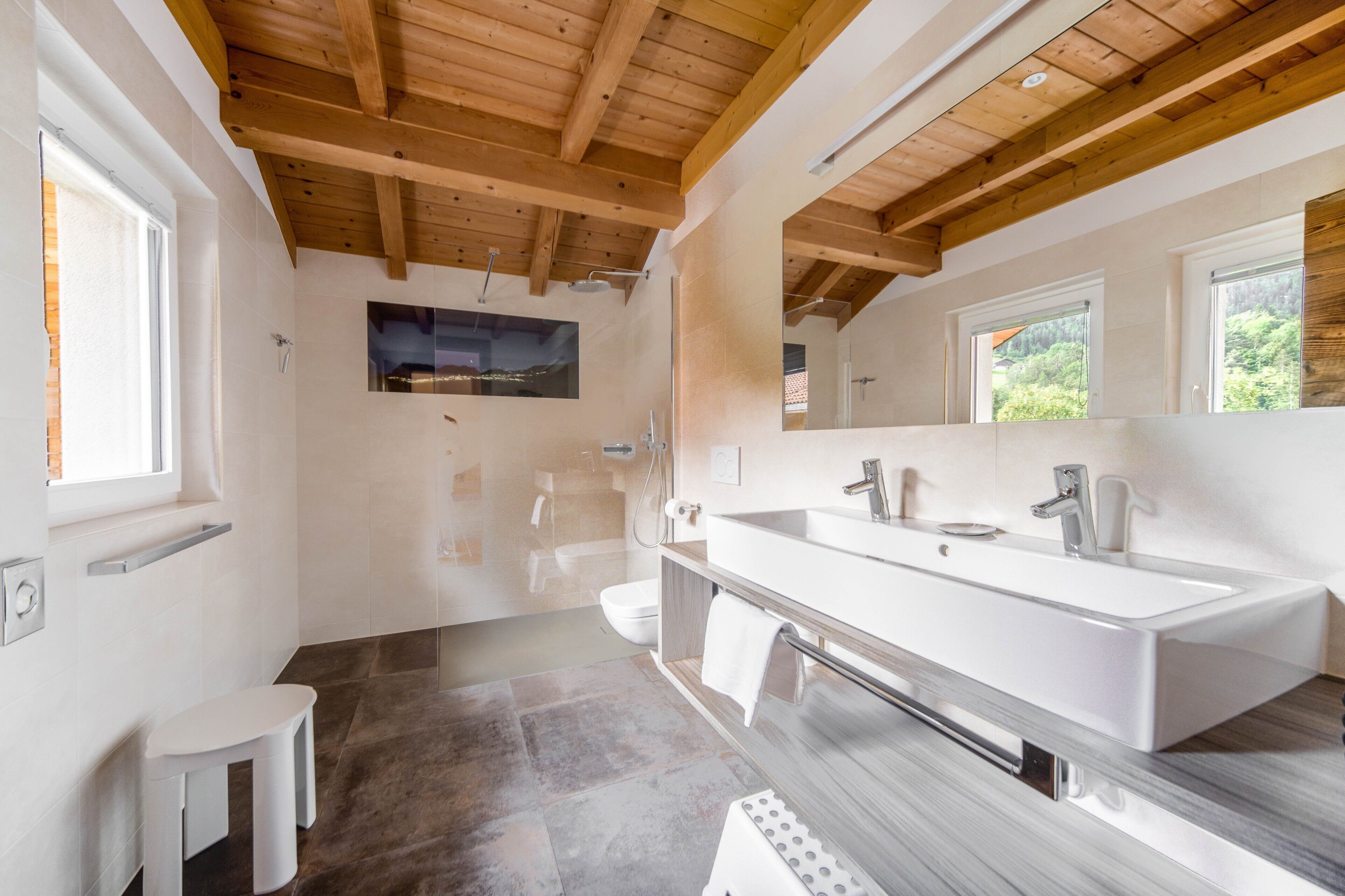 A modern bathroom with a large sink, a shower, and a small stool.