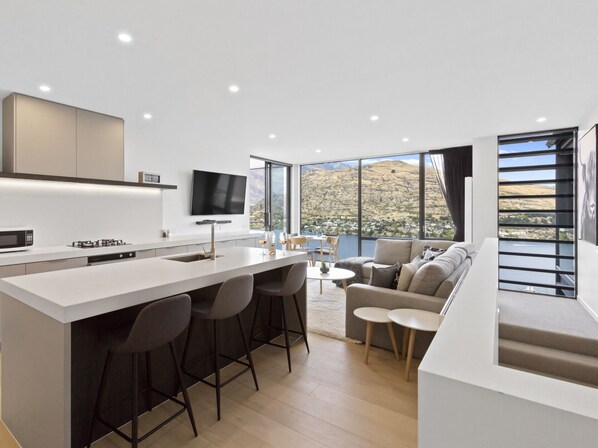 A modern kitchen with a central island, bar stools, a flat-screen TV, and large windows offering a scenic view. - Sierra Views - Queenstown Holiday Home (Queenstown)