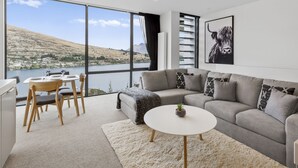 Ein modernes Wohnzimmer mit großem Fenster mit Blick auf einen See und Hügel, ein graues Sofa, ein kleiner runder Tisch und ein Essbereich mit Stühlen.